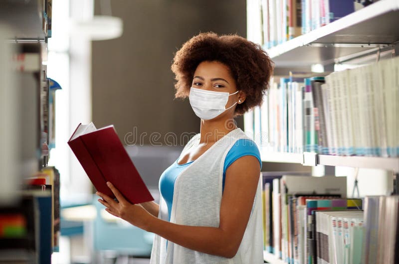 Studentessa africana in maschera con un libro in biblioteca immagine stock
