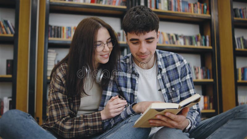 Studenti che studiano insieme in una biblioteca usando un portatile e prenotano su un pavimento intagliato circondato da scaffali video d archivio