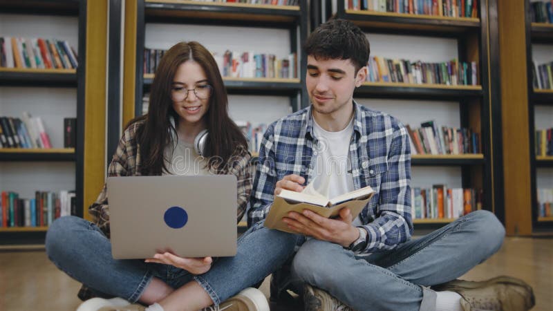 Studenti universitari studiano insieme in una biblioteca con un portatile e un libro seduti per terra a godersi la sessione di stu video d archivio