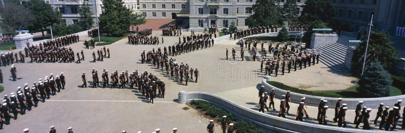 These are students of the United States Naval Academy marching in their noon meal formation. They are marching outdoors around a courtyard in their navy uniforms and white hats.