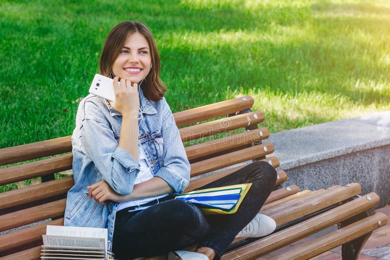 Studentă stă pe o bancă în parc și ține carnete și telefoane mobile. o fată învață lecții stock photography