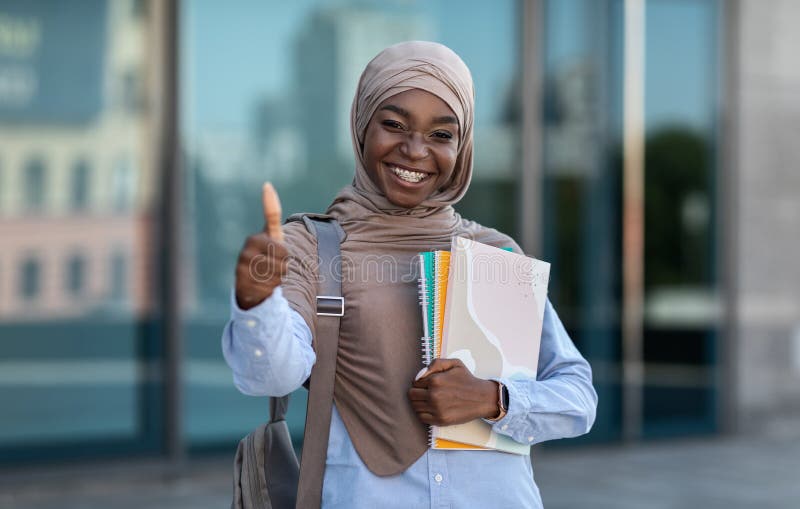 Study Programs For Islamic Students. Black Muslim Woman In Hijab Posing Outdoors, Showing Thumb Up, Smiling And Looking At Camera, Carrying Workbooks And Backpack, Resting At Campus After Classes. Study Programs For Islamic Students. Black Muslim Woman In Hijab Posing Outdoors, Showing Thumb Up, Smiling And Looking At Camera, Carrying Workbooks And Backpack, Resting At Campus After Classes