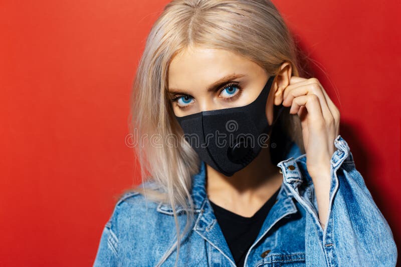 Studio close-up portrait of young blonde girl with blue eyes, wearing respiratory face mask of black color, against coronavirus.