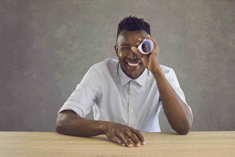 Happy black man sitting at desk and looking through hole in paper roll like in telescope royalty free stock photography