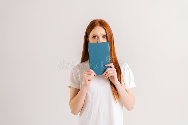 Studio portrait of scared young student woman hides face behind notebook looking at camera, standing on white isolated