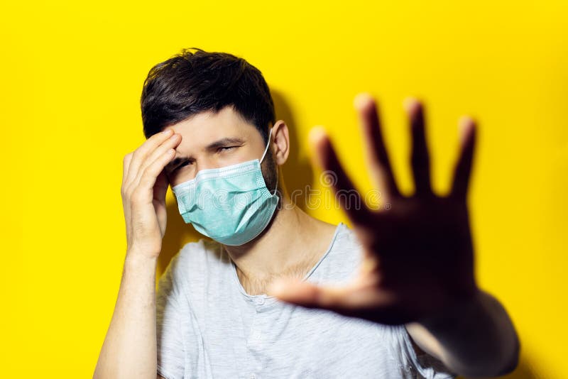 Studio- porträtt av ung sjuk man med huvudvärk, med mask för medicinsk influensa, visar gestalt STOP med hand mot bakgrund av gul royaltyfria bilder Studio- porträtt av ung sjuk man med huvudvärk, med mask för medicinsk influensa, visar gestalt STOP med hand mot bakgrund av gul royaltyfria bilder