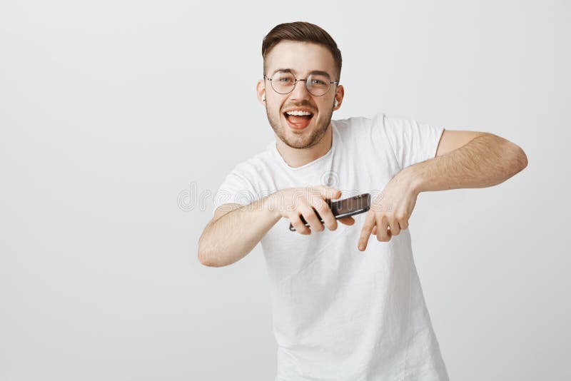 Studio shot of happy handsome and carefree young male model in white t-shirt singing along favorite song making rap moves near chest holding smartphone and dancing listening music in wireless earbuds.