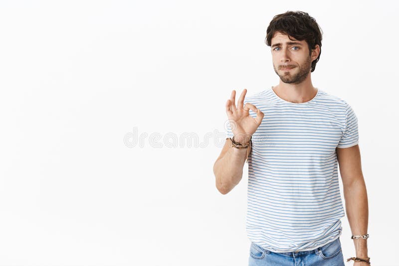 Studio shot of pressured and tired unsure young european male coworker showing okay gesture while pursing lips feeling
