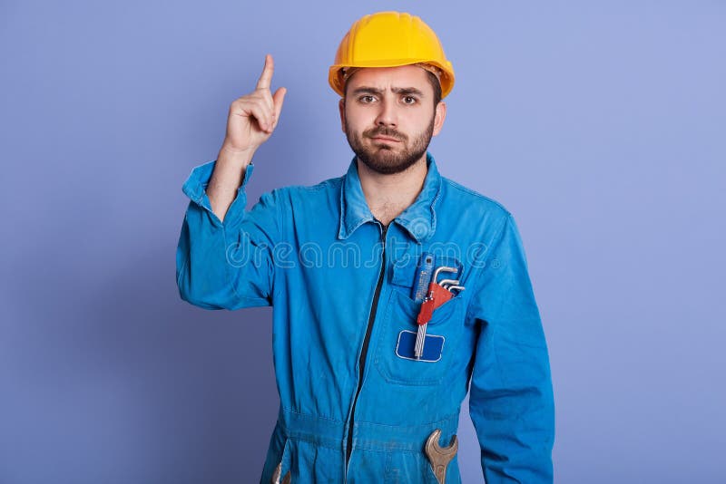 Studio shot of young construction worker with tools in pocket, dresses yellow helmet and uniform pointing up, bearded male having stock images Studio shot of young construction worker with tools in pocket, dresses yellow helmet and uniform pointing up, bearded male having stock images