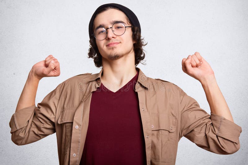 Studio shot of young man in cap, maroom t shirt, brown jacket and spectacles showing muscles. Isolated on a white background