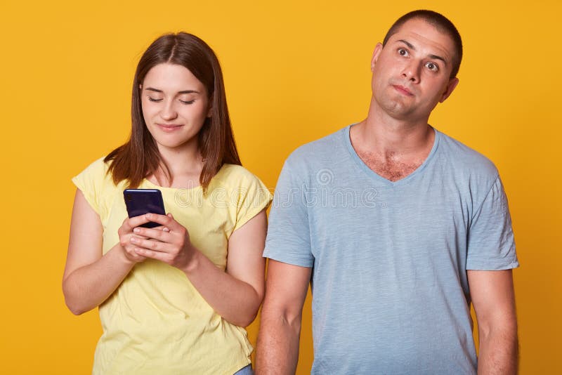 Studio shot of young couple, attractive female texting message on smartphone, male with thoughtful facial expression looking up, women and men wearing casually, posing isolated over yellow studio wall. Studio shot of young couple, attractive female texting message on smartphone, male with thoughtful facial expression looking up, women and men wearing casually, posing isolated over yellow studio wall