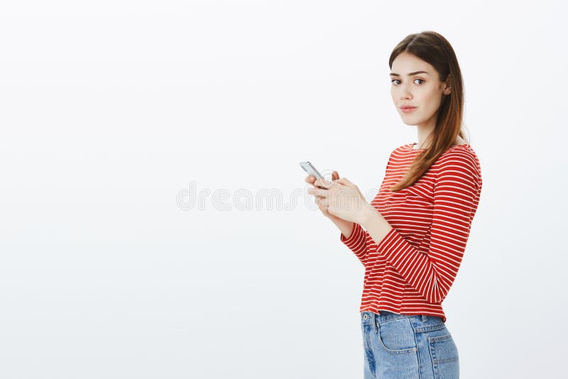 Studio portrait of gorgeous feminine european woman with brown hair, standing in profile, holding smartphone and looking at camera with casual, confident expression over gray background. Studio portrait of gorgeous feminine european woman with brown hair, standing in profile, holding smartphone and looking at camera with casual, confident expression over gray background.