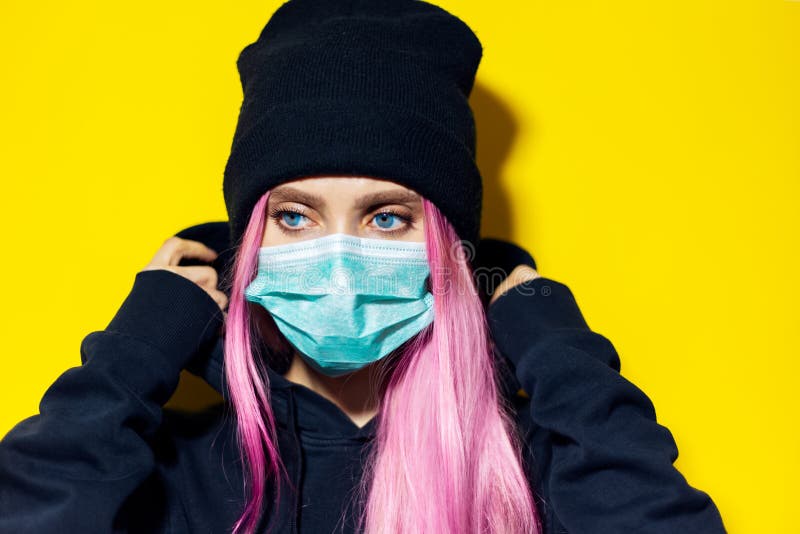 Studio portrait of young girl with pink hair and blue eyes, wearing medical flu mask, dressed in black hoodie sweater and beanie hat on yellow background. Studio portrait of young girl with pink hair and blue eyes, wearing medical flu mask, dressed in black hoodie sweater and beanie hat on yellow background