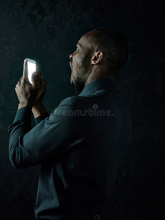 Studioskott av den unga allvarliga svarta afrikanska mannen som tänker, medan tala på mobiltelefonen mot svart bakgrund arkivfoto Studioskott av den unga allvarliga svarta afrikanska mannen som tänker, medan tala på mobiltelefonen mot svart bakgrund arkivfoto