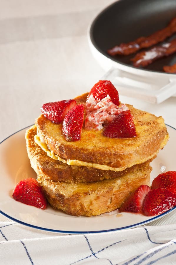 Organic stuffed french toast with fresh sliced strawberries on a blue rimed round white plate with strawberry butter and a span with three slices of fried bacon, all on a white table cloth with blue stripes. Organic stuffed french toast with fresh sliced strawberries on a blue rimed round white plate with strawberry butter and a span with three slices of fried bacon, all on a white table cloth with blue stripes