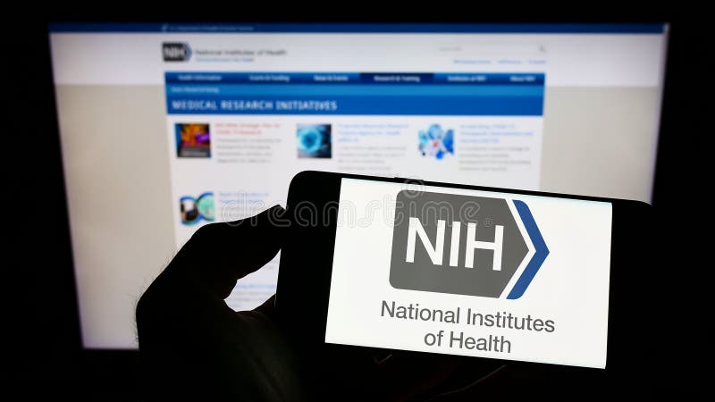 Person holding cellphone with logo of American agency National Institutes of Health (NIH) on screen in front of webpage. royalty free stock photos Person holding cellphone with logo of American agency National Institutes of Health (NIH) on screen in front of webpage. royalty free stock photos