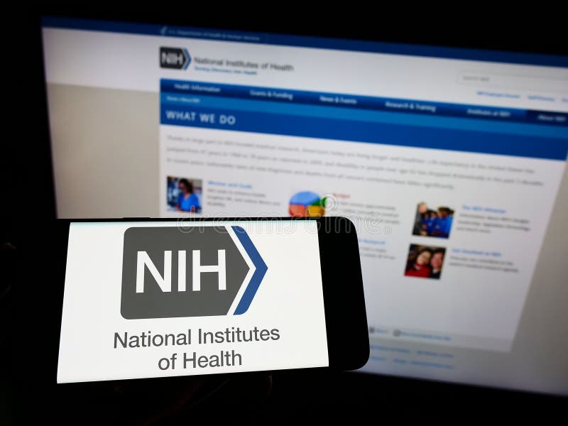 Person holding mobile phone with logo of US agency National Institutes of Health (NIH) on screen in front of web page. stock photo Person holding mobile phone with logo of US agency National Institutes of Health (NIH) on screen in front of web page. stock photo