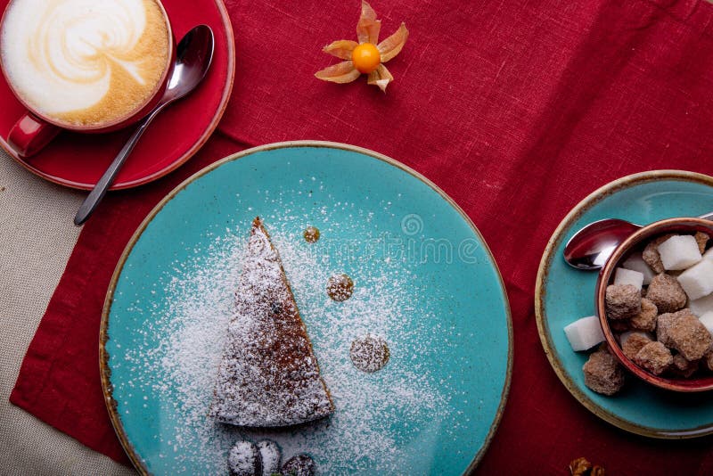 Stycke av morotkakan som täckas med pudrat socker i en blå platta på en röd servett med koppen av coffe och farin arkivfoto Stycke av morotkakan som täckas med pudrat socker i en blå platta på en röd servett med koppen av coffe och farin arkivfoto