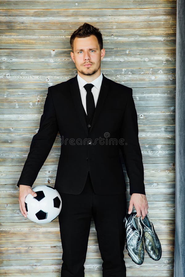 Stylish athletic man in a business suit holding a soccer ball with football boots. Against the background of a loft wall. Fashionable hairstyle and black tie. Coach or football player. Self-confident. Stylish athletic man in a business suit holding a soccer ball with football boots. Against the background of a loft wall. Fashionable hairstyle and black tie. Coach or football player. Self-confident.