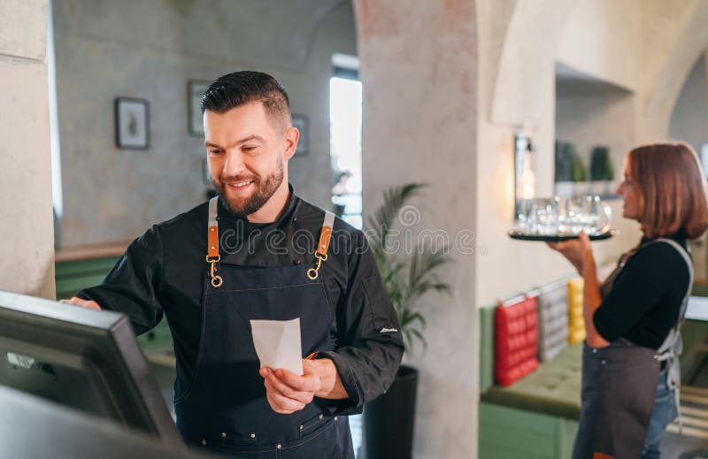 Stylish bearded smiling waiter dressed black uniform using point of sale order terminal system touch Screen and waitress woman stock image