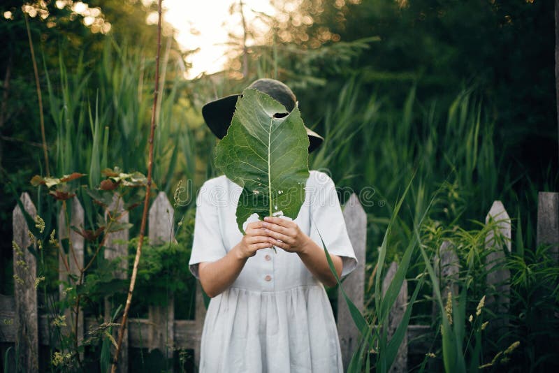 Stylish girl in linen dress holding big green leaf at face at wooden fence and grass. Portrait of boho woman in hat posing with