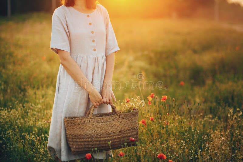 Stylish girl in linen dress holding rustic straw basket with poppy flowers in meadow in sunset light. Boho woman relaxing and royalty free stock photo Stylish girl in linen dress holding rustic straw basket with poppy flowers in meadow in sunset light. Boho woman relaxing and royalty free stock photo
