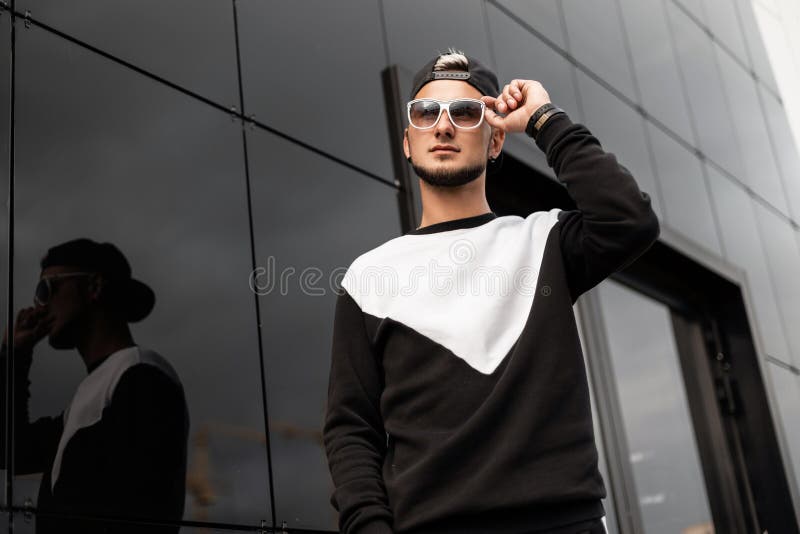Stylish handsome young hipster man in trendy white sunglasses in fashionable sweatshirt in American black cap poses royalty free stock photography Stylish handsome young hipster man in trendy white sunglasses in fashionable sweatshirt in American black cap poses royalty free stock photography