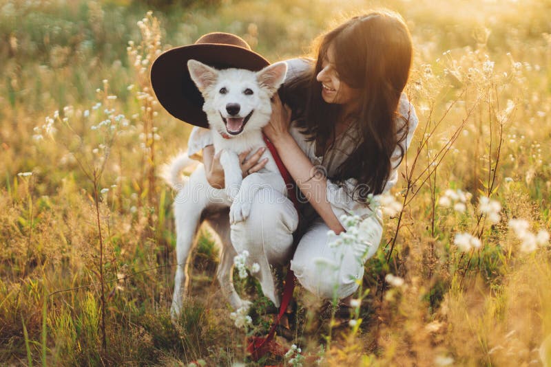 Stylish happy woman playing with cute dog with hat among wildflowers in sunset light. Summer travel with pet. Young carefree