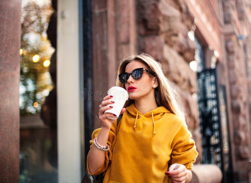 Stylish happy young woman wearing boyfrend jeans, white sneakers bright yellow sweatshirt.She holds coffee to go. portrait of smil