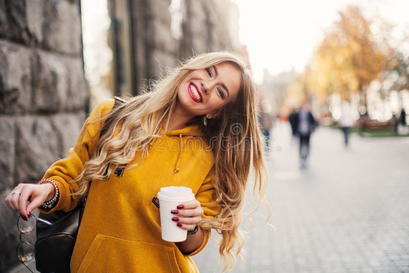 Stylish happy young woman wearing boyfrend jeans, white sneakers bright yellow sweetshot.She holds coffee to go. portrait of smili