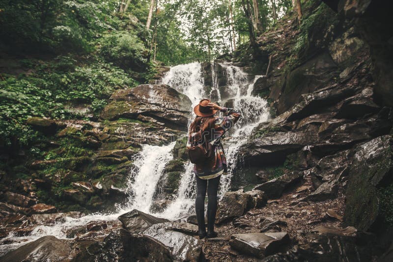 Stylish hipster girl in hat traveling in mountains. Young woman with backpack exploring in summer forest, looking at waterfall. stock photo Stylish hipster girl in hat traveling in mountains. Young woman with backpack exploring in summer forest, looking at waterfall. stock photo