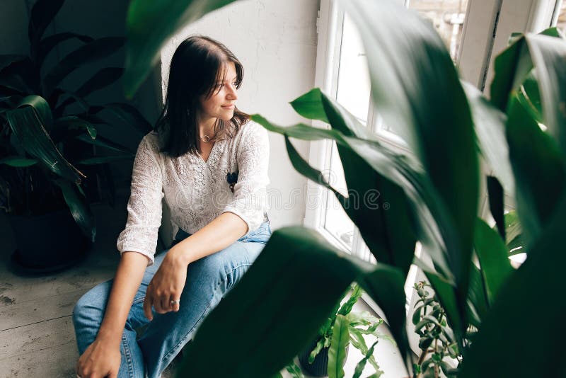 Stylish hipster girl sitting on floor and looking at white rustic window with green plants in modern cafe. Fashionable woman