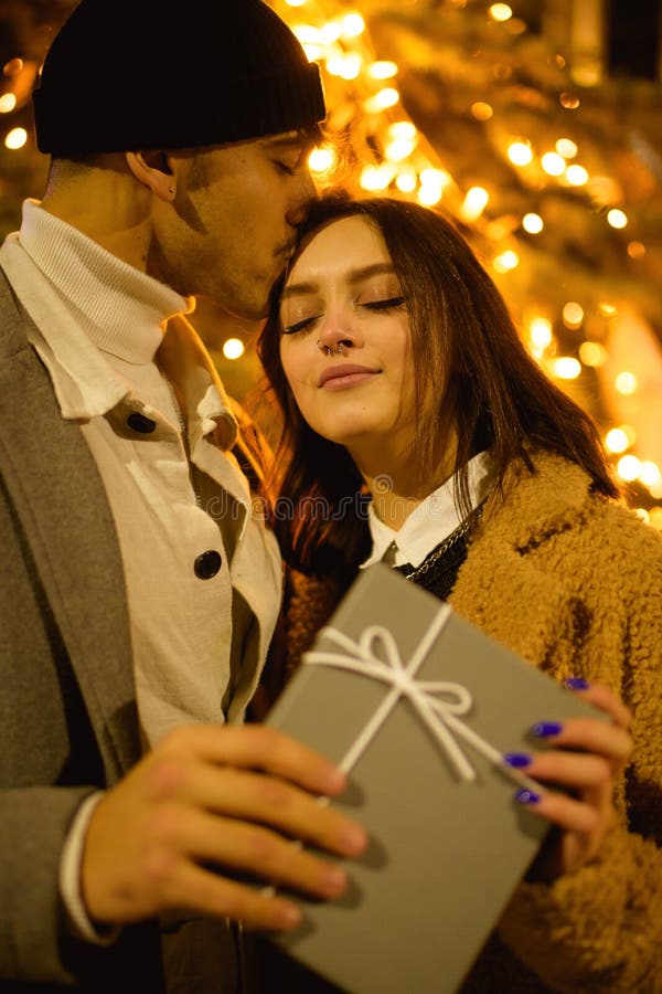 Stylish Man Kissing Girlfriend With Present Near Garland In Evening