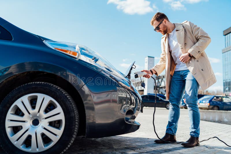 Stylish man in sunglasses inserts the charging cable into the socket of electric car.