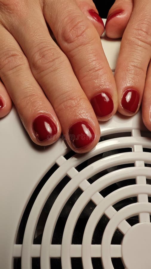 Hands adorned with a glossy red manicure rest on a salon dryer, the warm atmosphere highlighting the care and attention given to nail artistry during a relaxing evening. Pampering experience stock images, royalty-free photos and pictures