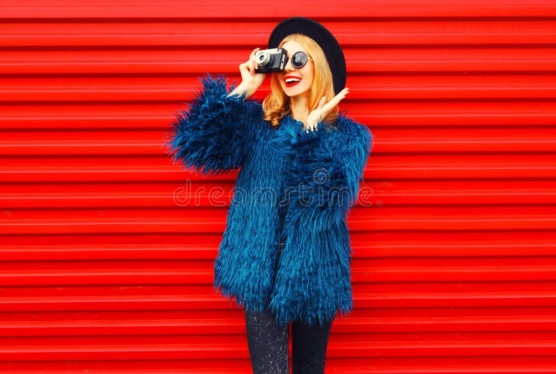 Stylish surprised woman with retro camera taking picture wearing blue faux fur coat, round hat and sunglasses over colorful red stock images Stylish surprised woman with retro camera taking picture wearing blue faux fur coat, round hat and sunglasses over colorful red stock images
