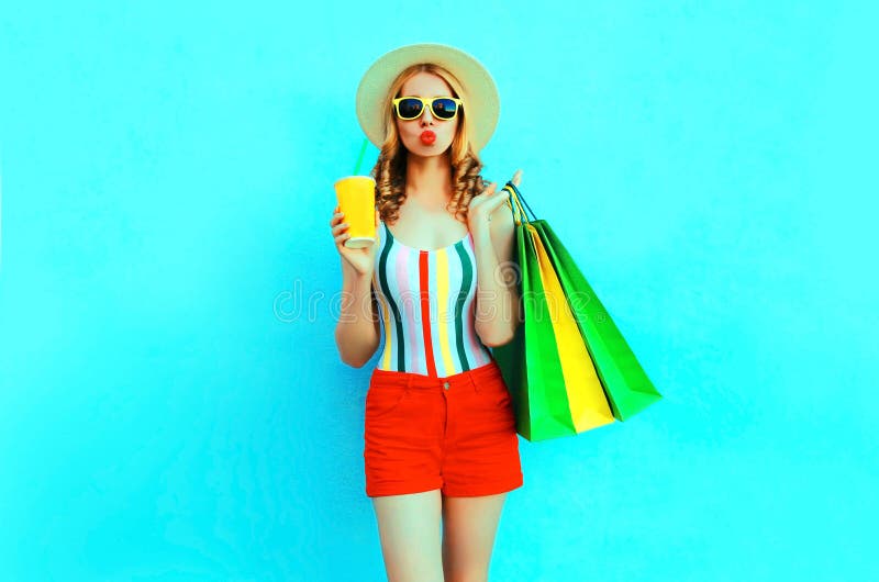 Stylish woman holding cup of fruit juice with shopping bags in colorful t-shirt, summer straw hat, sunglasses, red shorts on blue. Background stock photo Stylish woman holding cup of fruit juice with shopping bags in colorful t-shirt, summer straw hat, sunglasses, red shorts on blue. Background stock photo