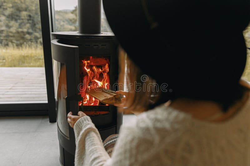 Stylish woman in knitted sweater putting firewood in to modern black fireplace with warm fire