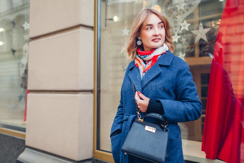 Stylish woman wearing scarf blue coat holding purse by shop on street. Spring fashion female accessories and clothes.