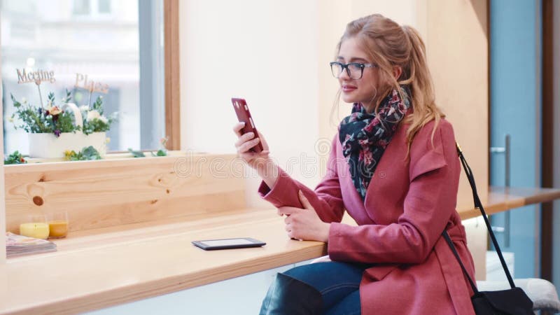 Stylish young European girl with curly hairstyle happily smiling while using her cellphone for texting the message