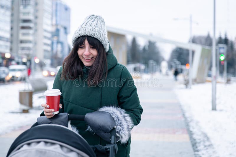 Stylish young mom with a cup of coffee on a walk with a baby carriage in winter.