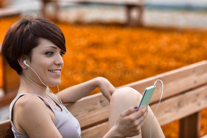 Stylish young woman sitting on a bench listening to music with a blissful smile of pleasure in a close up side view. Stylish young woman sitting on a bench listening to music with a blissful smile of pleasure in a close up side view