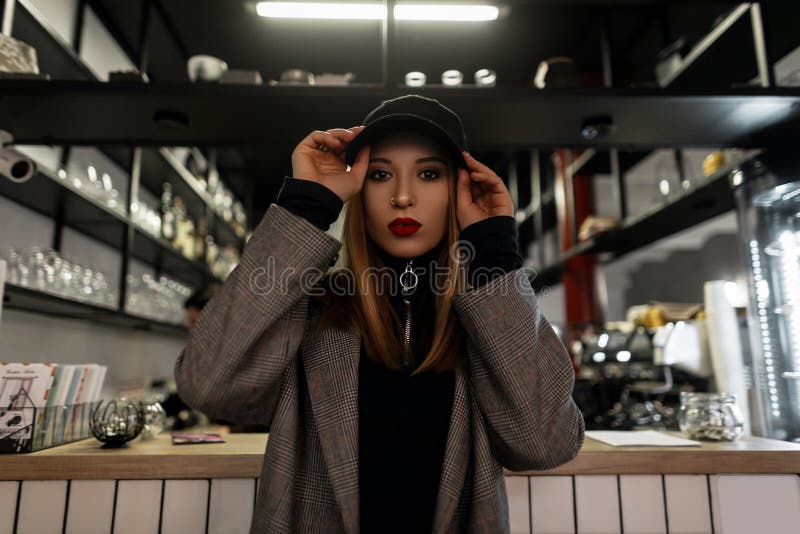Stylowa młoda kobieta z przekłutym nosem w modnej czarnej czapce w szarej, zabytkowej, kieszonkowej kurtce z czerwonymi ustami po fotografia stock