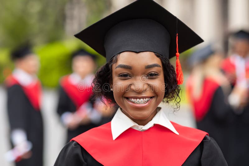 Closeup portrait of happy young black lady student wearing graduating costume. Happy african american female student celebrating graduation with her friends at university campus, blurred background. Closeup portrait of happy young black lady student wearing graduating costume. Happy african american female student celebrating graduation with her friends at university campus, blurred background