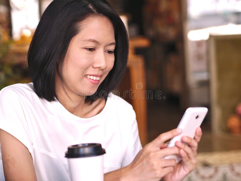 Stående av den asiatiska kvinnan som skriver till hennes smartphoneskärm med den lyckliga framsidan för leende i hemtrevlig coffe arkivbild Stående av den asiatiska kvinnan som skriver till hennes smartphoneskärm med den lyckliga framsidan för leende i hemtrevlig coffe arkivbild