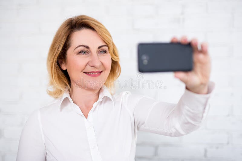 Stående av den gladlynta kvinnan för mogen affär som tar selfie med den smarta telefonen över den vita väggen arkivbild Stående av den gladlynta kvinnan för mogen affär som tar selfie med den smarta telefonen över den vita väggen arkivbild