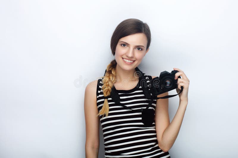 Stående av den lyckliga unga härliga kvinnan i den randiga skjortan som poserar med den svarta fotokameran mot studiobakgrund arkivbild Stående av den lyckliga unga härliga kvinnan i den randiga skjortan som poserar med den svarta fotokameran mot studiobakgrund arkivbild