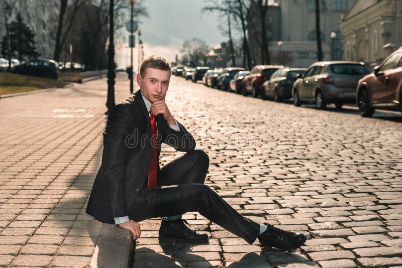 Stående av en brutal man i en svart dräkt och ett rött band En ung grabb sitter på gatan längs det gammalt stenar vägen arkivfoton Stående av en brutal man i en svart dräkt och ett rött band En ung grabb sitter på gatan längs det gammalt stenar vägen arkivfoton