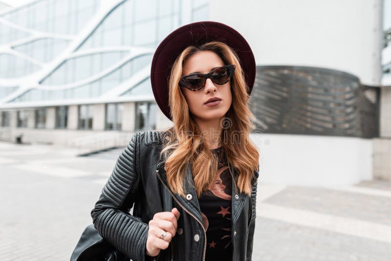 Stående av en redhaired attraktiv ung hipsterkvinna i en elegant hatt i moderiktig solglasögon i ett svartläderomslag royaltyfria bilder Stående av en redhaired attraktiv ung hipsterkvinna i en elegant hatt i moderiktig solglasögon i ett svartläderomslag royaltyfria bilder