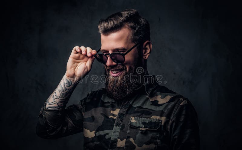 Stående av en stilfull skäggig grabb med tatuerade händer i den militära skjortainnehavhanden på solglasögon och skratt arkivfoto Stående av en stilfull skäggig grabb med tatuerade händer i den militära skjortainnehavhanden på solglasögon och skratt arkivfoto
